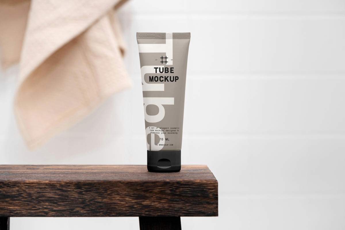 Front view, a Cosmetic Tube Mockup with delicate cosmetic design in black, cream, and beige colors, standing on a wooden stool, which is standing in the brightly lit bathroom.