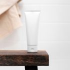 Front view, a white Cosmetic Tube Mockup standing on a wooden stool, which is standing in the brightly lit bathroom.