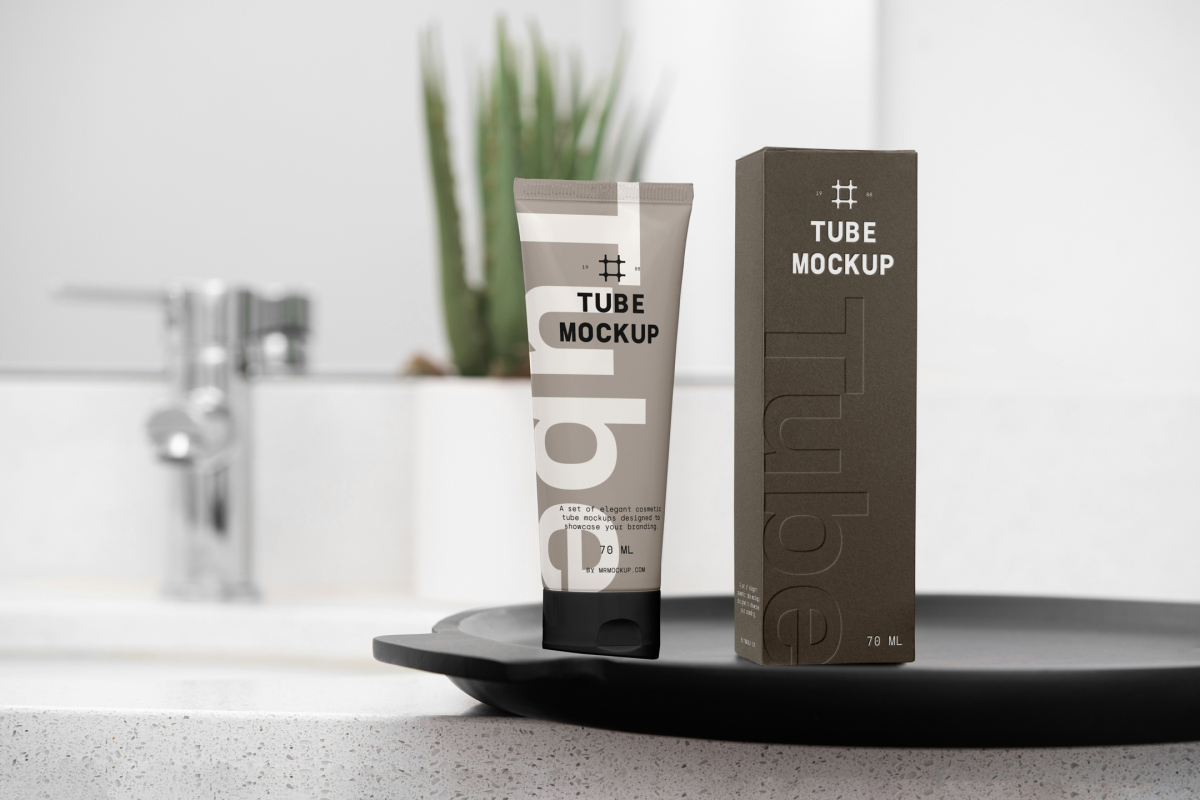 Front view, a Cosmetic Tube Mockup and a white Box Mockup with delicate cosmetic designs in black, cream, beige, and dark green colors, stand next to each other on a black colored tray. In the background you can see a brightly lit bathroom.