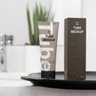Front view, a Cosmetic Tube Mockup and a white Box Mockup with delicate cosmetic designs in black, cream, beige, and dark green colors, stand next to each other on a black colored tray. In the background you can see a brightly lit bathroom.