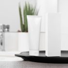 Front view, a white Cosmetic Tube Mockup and a white Box Mockup stand next to each other on a black colored tray. In the background you can see a brightly lit bathroom.