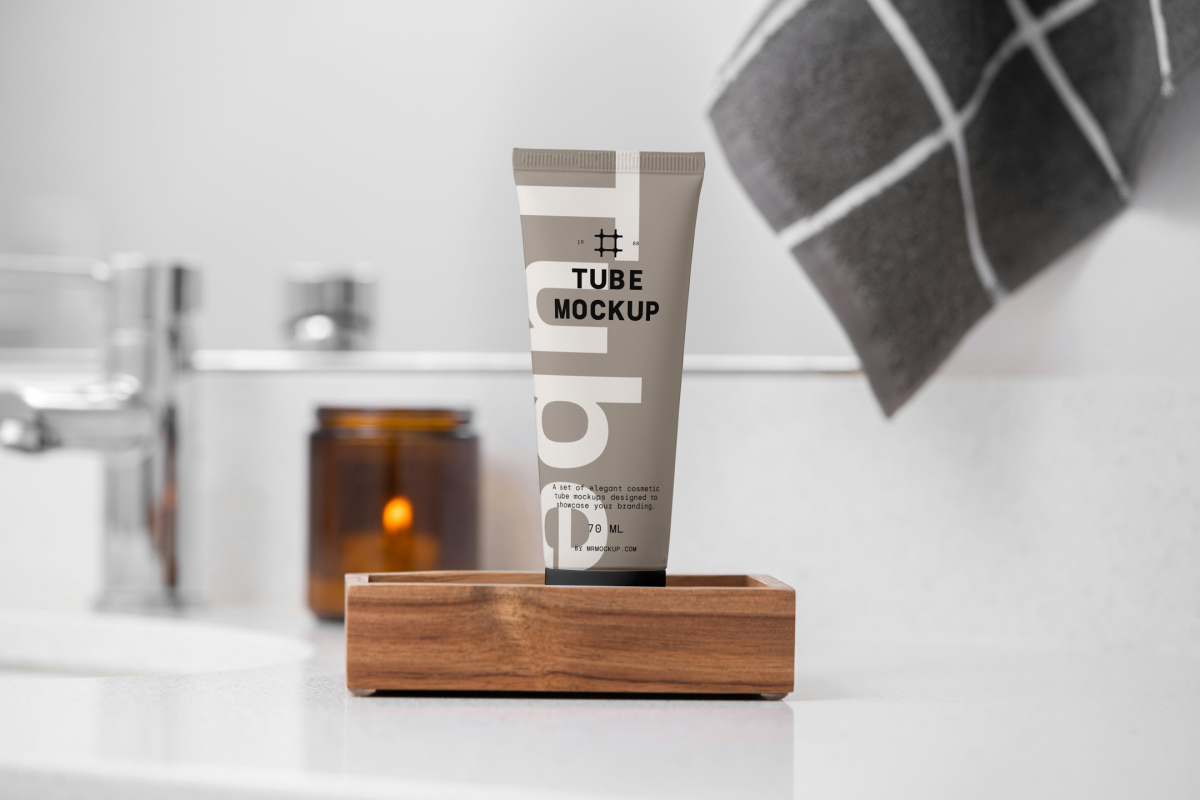 Front view, a Cosmetic Tube Mockup with delicate cosmetic design in black, cream, and beige colors, standing in the wooden box, which lies on a white sink. In the background you can see a brightly lit bathroom.