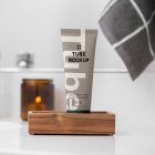 Front view, a Cosmetic Tube Mockup with delicate cosmetic design in black, cream, and beige colors, standing in the wooden box, which lies on a white sink. In the background you can see a brightly lit bathroom.