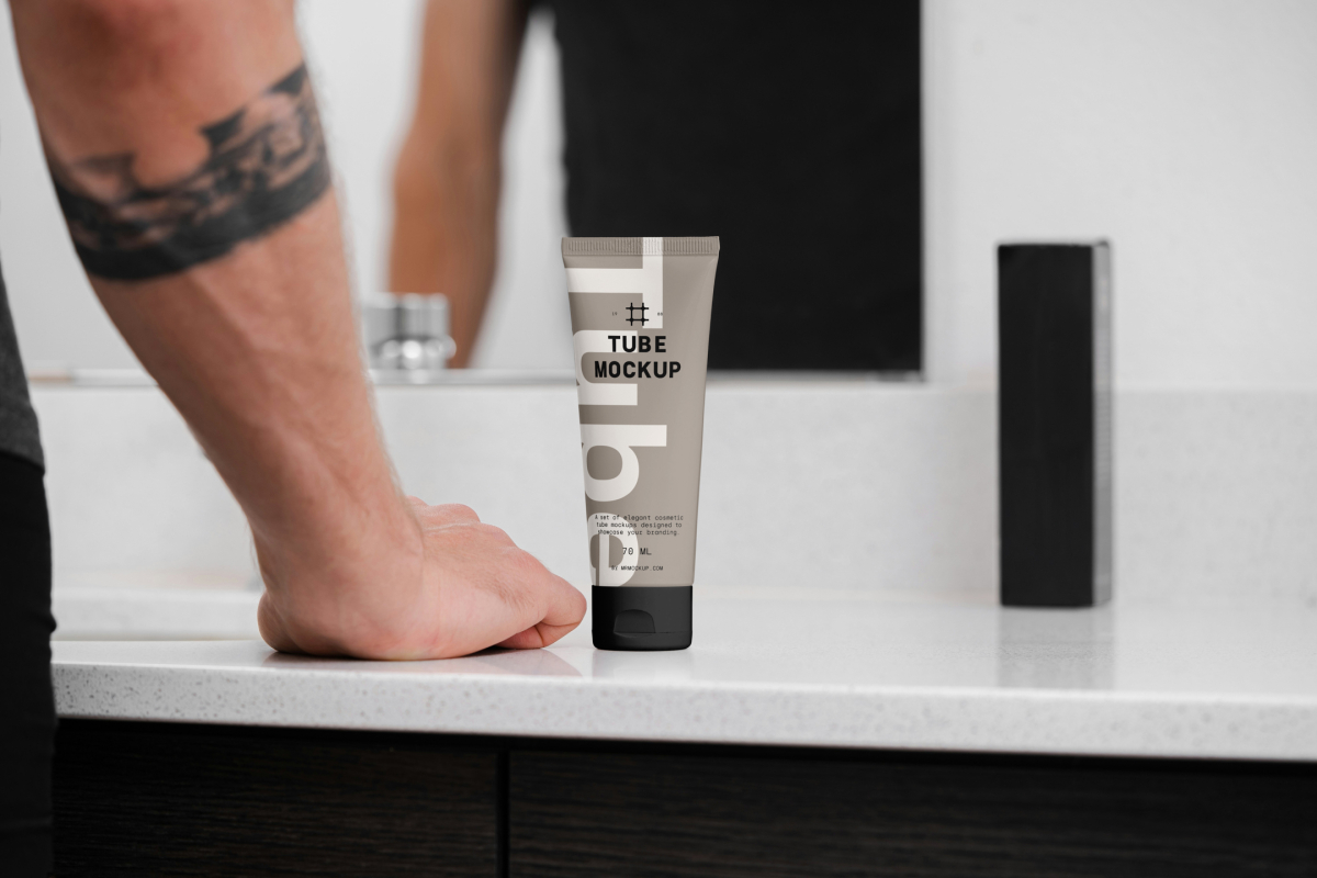 Front view, a Cosmetic Tube Mockup with delicate cosmetic design in black, cream, and beige colors, standing on a bright color marble sink. In the background you can see a brightly lit bathroom and men standing in front of the mirror.