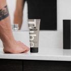 Front view, a Cosmetic Tube Mockup with delicate cosmetic design in black, cream, and beige colors, standing on a bright color marble sink. In the background you can see a brightly lit bathroom and men standing in front of the mirror.
