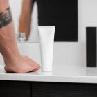 Front view, a white Cosmetic Tube Mockup standing on a bright color marble sink. In the background you can see a brightly lit bathroom and standing men in front of the mirror.