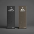 In the front view, two vertical boxes from the Cosmetic Tube Mockups product with delicate cosmetic designs in black, dark green, and white colors stand next to each other on the dark background.