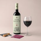 Front view, the scene from Studio Wine Mockups of a standing black wine bottle with a green label, a transparent wine glass with typography design, and two paper pads in dark gray and claret colors on the pink background.