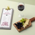 View from above, scene from Studio Wine Mockups wooden menu with a typography design, standing transparent wine glass with a typography design, and lying wooden tray with camembert and grapes on the bright green background.