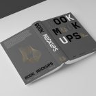 View from above, an open hardcover book mockup turned upside down with bold typography design in gold, black, gray, and white colors lying on the white background in perspective view.