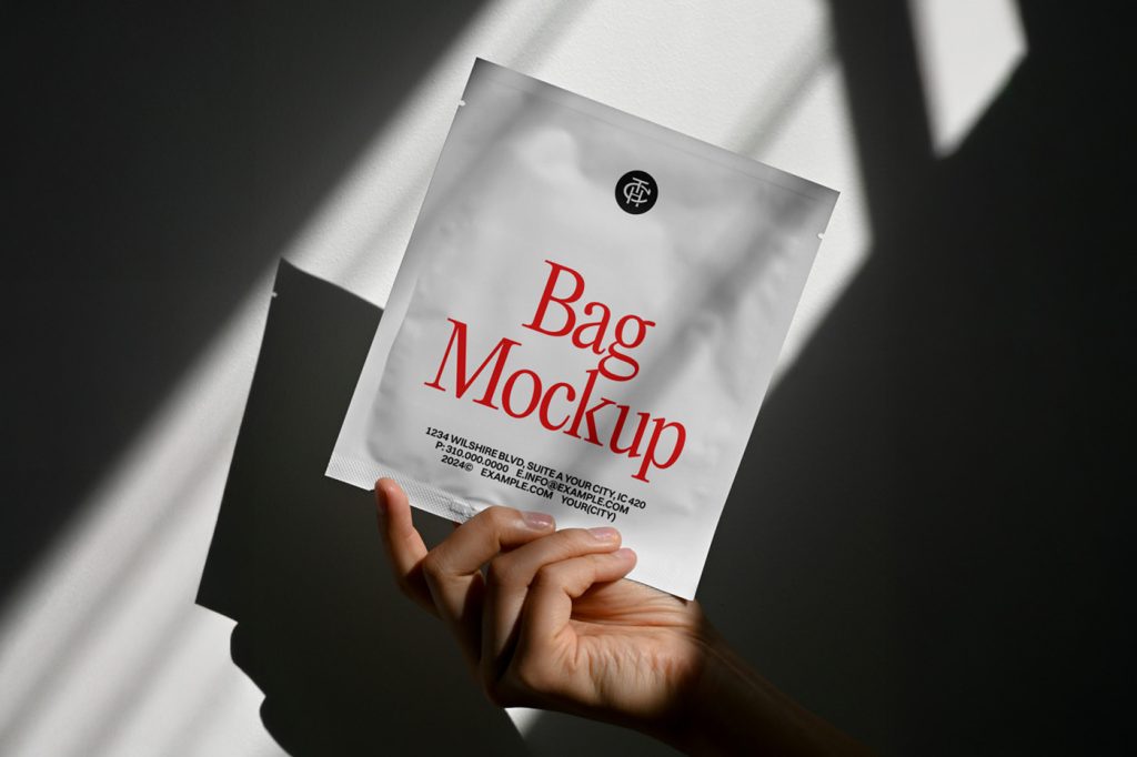 Front view, hand holding white bag mockup with red and black typography design against the white wall with shadows.
