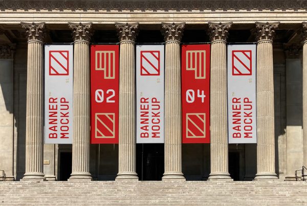 Front view, five vertical mockups with white, red, and cream geometric designs hanging between the columns on the facade of the building just above the entrance to the museum.