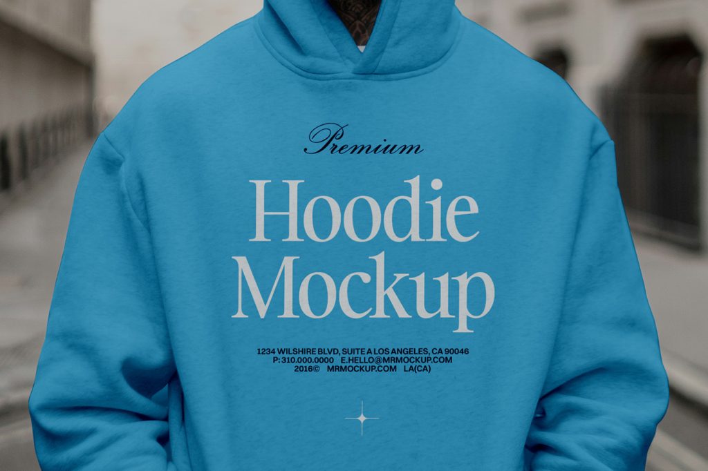 Front view, standing male model wearing a baby blue hoodie color mockup with a serif typography design against the city view.