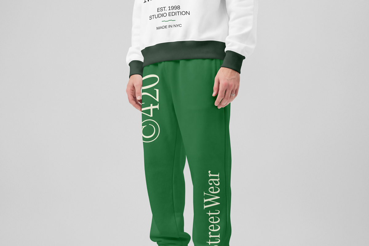 Front view of a standing male model wearing green sweatpants & a white sweatshirt mockup with a typography design against a grey studio background from the studio apparel mockups product.