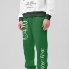 Front view of a standing male model wearing green sweatpants & a white sweatshirt mockup with a typography design against a grey studio background from the studio apparel mockups product.