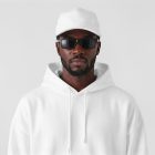 Front view of a Black male model wearing a white cap & a white hoodie mockup standing against the white studio background from the studio apparel mockups product.