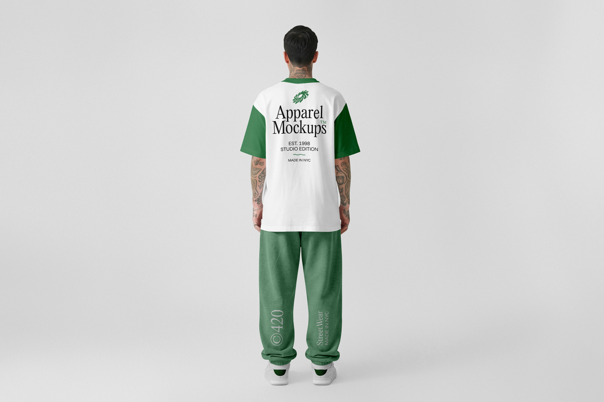 Front view, male model standing backward wearing a white t-shirt & a pastel green sweatpants mockup with typography design against a grey studio background from the studio apparel mockups product.