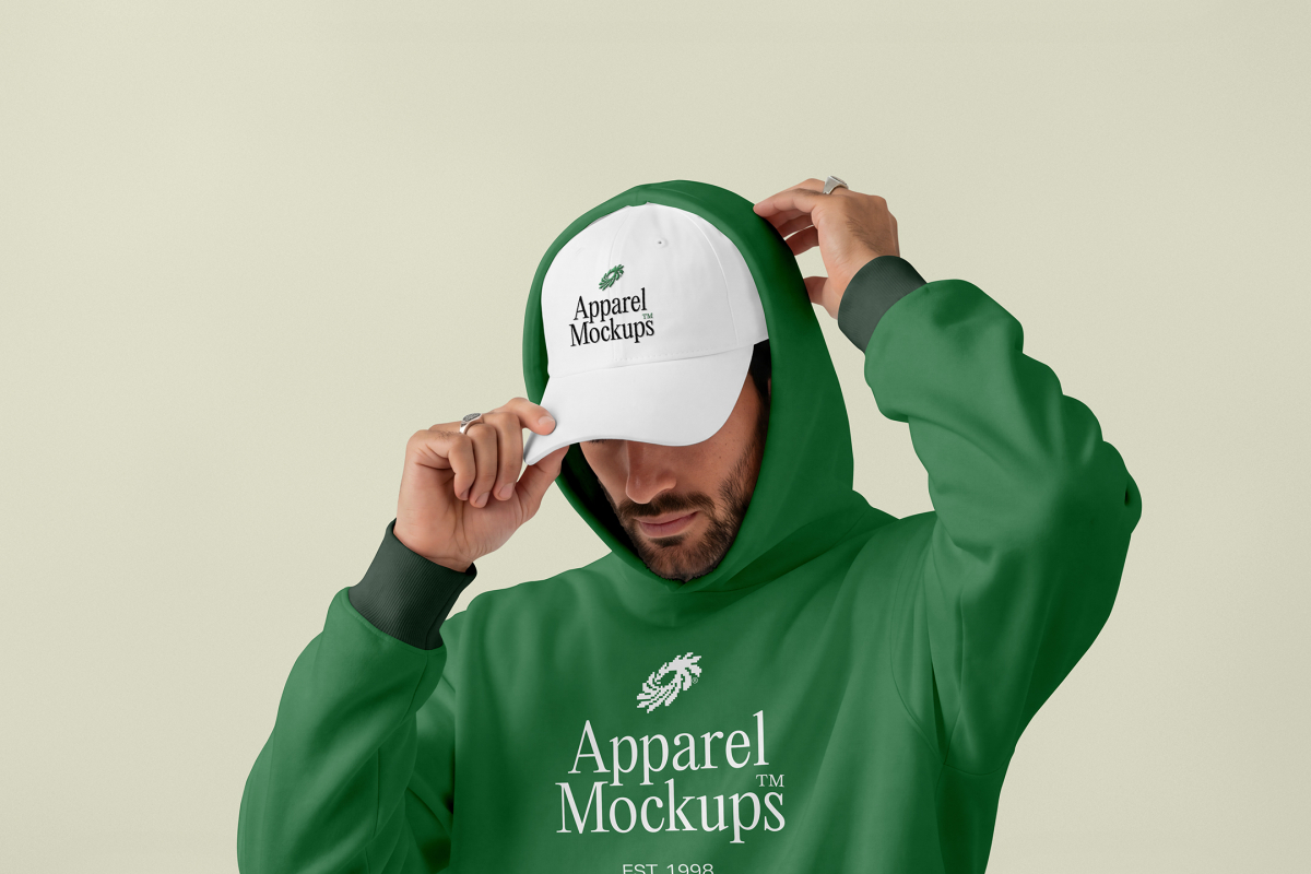 Front view, male model wearing a white cap & a green hoodie mockup with a typography design standing against the bright studio background from studio apparel mockups product.
