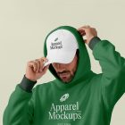 Front view, male model wearing a white cap & a green hoodie mockup with a typography design standing against the bright studio background from studio apparel mockups product.