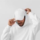 Front view, male model wearing a white cap & a white hoodie mockup standing against the white studio background from the studio apparel mockups product.