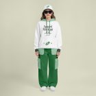 Front view of a female model wearing green baggy pants, a white hoodie, & a white cap mockup with a typography design standing against a bright studio background from the studio apparel mockups product.
