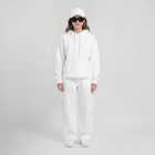 Front view of a female model wearing white baggy pants, a white hoodie, & a white cap mockup standing against a white studio background from the studio apparel mockups product.