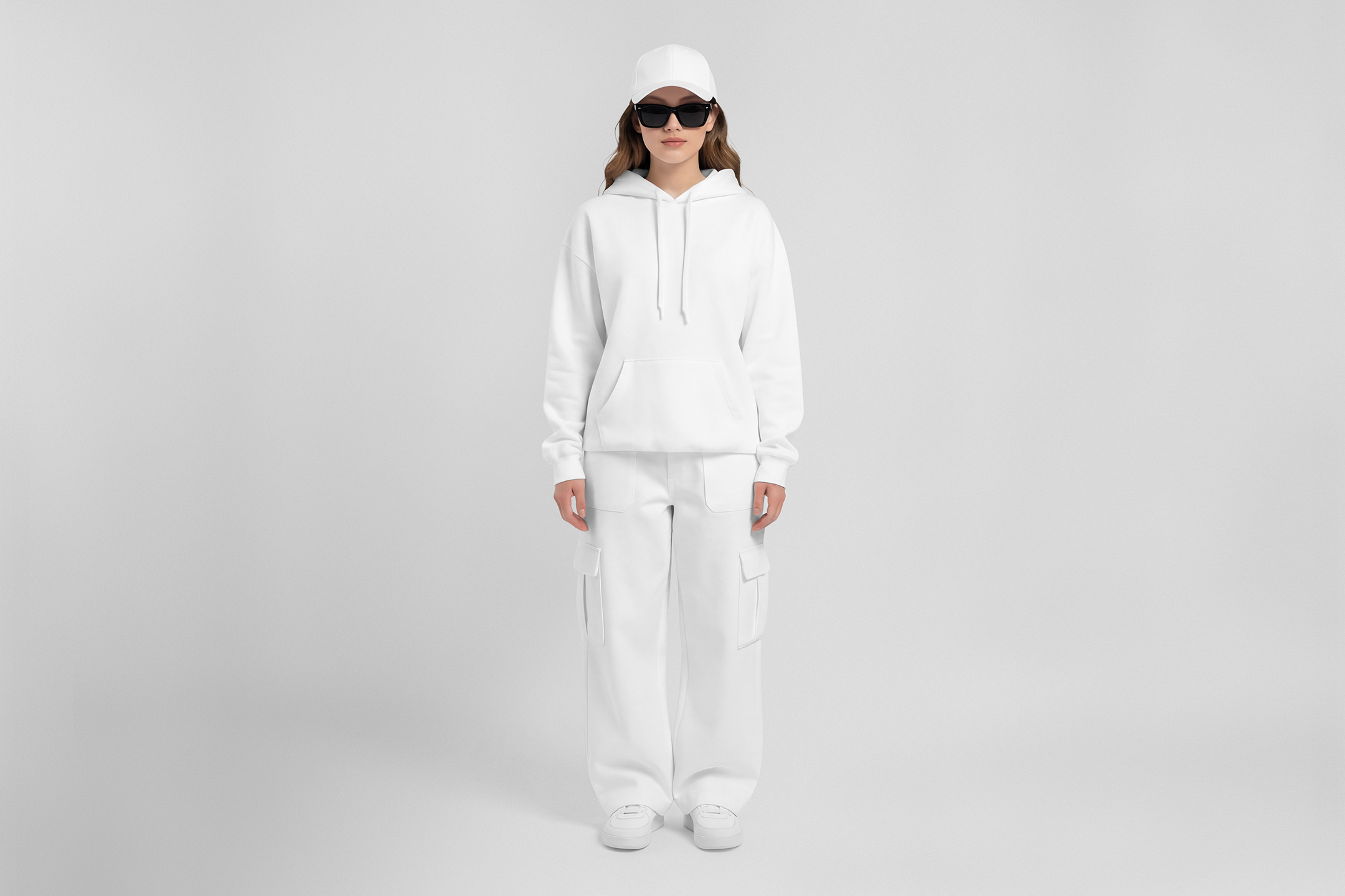 Front view of a female model wearing white baggy pants, a white hoodie, & a white cap mockup standing against a white studio background from the studio apparel mockups product.