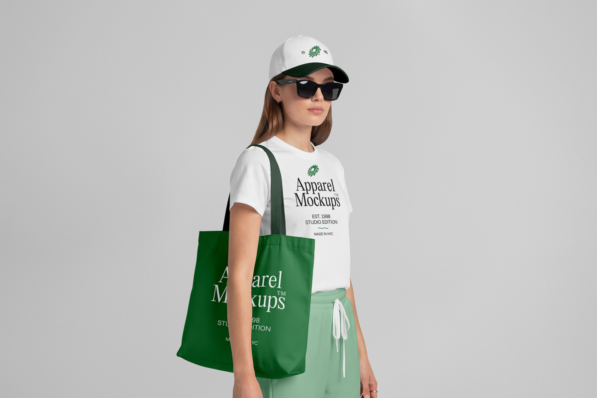 Front view of a female model wearing a green canvas bag, a white t-shirt, & a white cap mockup with a typography design standing against a grey studio background from the studio apparel mockups product.