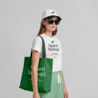 Front view of a female model wearing a green canvas bag, a white t-shirt, & a white cap mockup with a typography design standing against a grey studio background from the studio apparel mockups product.