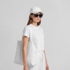 Front view of a female model wearing a white canvas bag, a white t-shirt, & a white cap mockup standing against a white studio background from the studio apparel mockups product.
