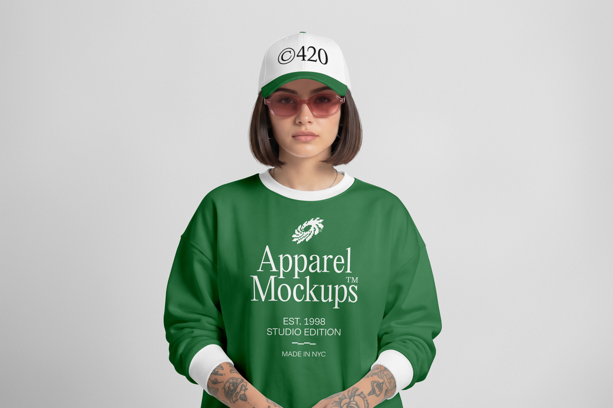 Front view of a female model wearing a green sweatshirt and white cap mockup with a white and black typography design standing against the grey studio background from the studio apparel mockups product.