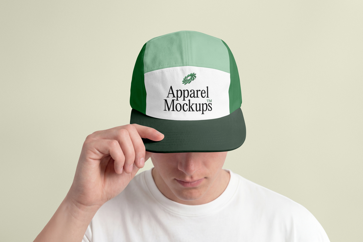 Front view of a male model wearing a five panel cap mockup in three shades of green and white colors with a black typography design held by hand, standing against the bright studio background from the studio apparel mockups product.
