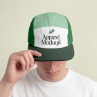 Front view of a male model wearing a five panel cap mockup in three shades of green and white colors with a black typography design held by hand, standing against the bright studio background from the studio apparel mockups product.