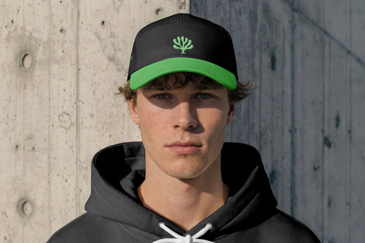 Front view, white male model wearing a hoodie mockup and cap mockup with a green logo and peak. Standing in front of a big concrete wall from the Outdoor Apparel Mockups Vol.3 product.