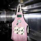A light pink kitchen apron hanging from a metal hook attached to a stainless steel industrial stove hood. The apron features a large green logo, patterned pockets, and several text stickers.
