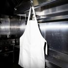A blank white cooking apron hanging from a metal hook attached to a stainless steel hood in a professional kitchen. A stove and pots are visible blurred in the background.