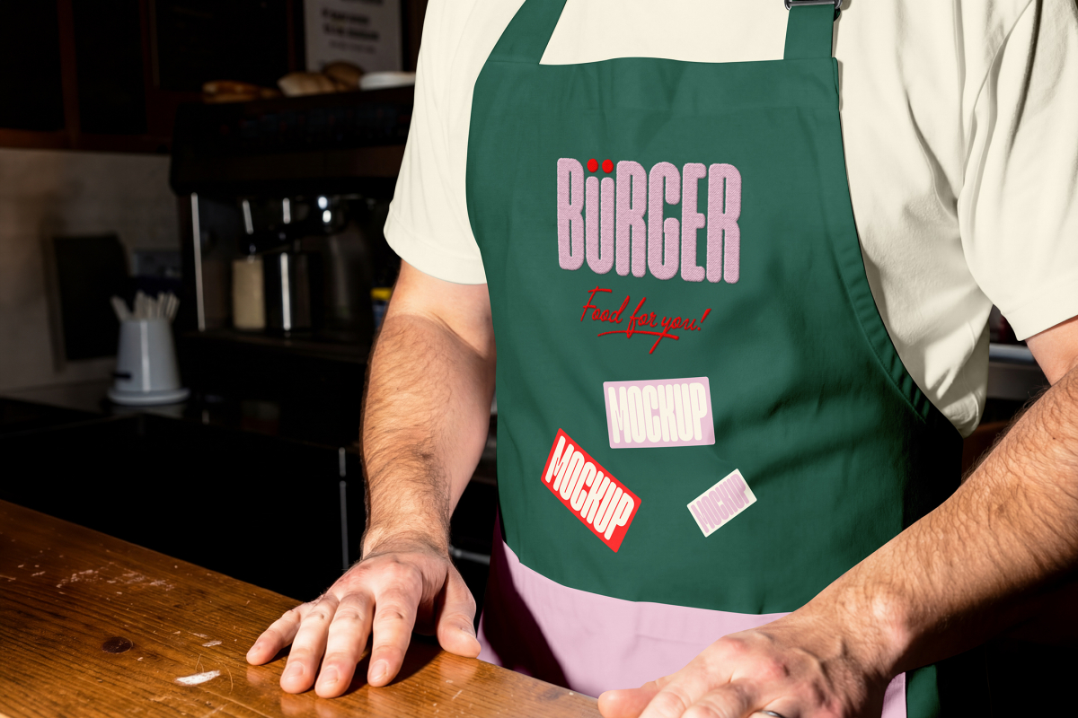 A close-up of a person standing behind a wooden counter, wearing a dark green apron over a cream-colored t-shirt. The apron features a pink embroidered "BÜRGER" logo and multiple rectangular mockup stickers.