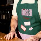 A close-up of a person standing behind a wooden counter, wearing a dark green apron over a cream-colored t-shirt. The apron features a pink embroidered "BÜRGER" logo and multiple rectangular mockup stickers.