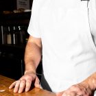 A close-up shot of a person standing behind a wooden counter wearing a plain white t-shirt and a matching white apron. The background features a blurred espresso machine.