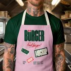 A close-up of a person with tattoos on their neck and arms, wearing a light pink apron over a dark green t-shirt in a professional food truck kitchen. The apron displays a large dark green brand logo and mockup labels.