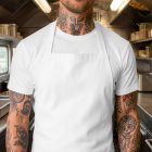 A close-up front view of a person with extensive tattoos on their neck and arms, wearing a plain white apron over a matching white t-shirt inside a stainless steel food truck kitchen.