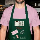 A close-up of a person wearing a dark green apron over a light pink t-shirt in a commercial kitchen setting. The apron is customized with a textured "BÜRGER" logo, red tagline, and mockup badge graphics.