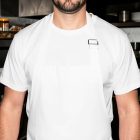A close-up of a bearded man from the chest down, wearing a plain white cooking apron over a blank white t-shirt in a professional kitchen setting.