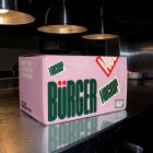 A large, rectangular pink delivery box resting on a brushed stainless steel kitchen counter under industrial warmth lamps. The side of the box shows prominent green branding and sticker graphics.