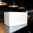 A blank white rectangular cardboard packaging box sitting on a brushed stainless steel kitchen counter under warm, industrial hanging lamps.