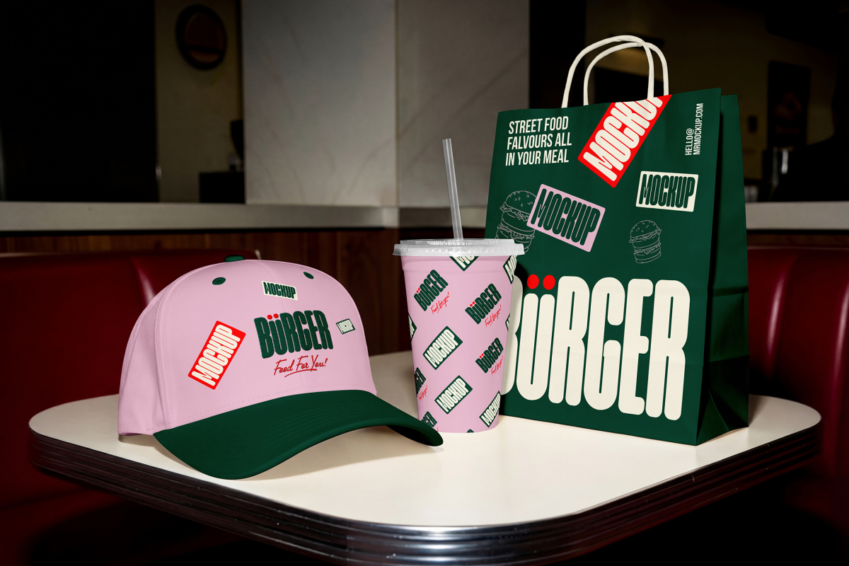 A branding presentation on a white diner table featuring a pink baseball cap with a dark green brim, a pink paper cup covered in a green logo pattern, and a dark green paper takeout bag with large white typography and mockup labels.