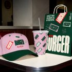 A branding presentation on a white diner table featuring a pink baseball cap with a dark green brim, a pink paper cup covered in a green logo pattern, and a dark green paper takeout bag with large white typography and mockup labels.