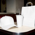 A fast-food branding mockup set arranged on a white diner table, featuring a plain white baseball cap, a blank white beverage cup with a clear lid and straw, and a blank white paper shopping bag with loop handles.