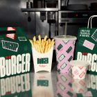 A collection of branded fast-food packaging lined up on a reflective metal counter, including a green take-out carton, a white fry box filled with golden french fries, a pink patterned drink cup, a small sauce container, and a green paper shopping bag.