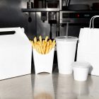 A selection of blank white fast-food packaging lined up on a reflective metal counter, including a gable-top box with a handle, a fry scoop filled with french fries, a large plastic-lidded drink cup, a small sauce cup, and a paper take-out bag.